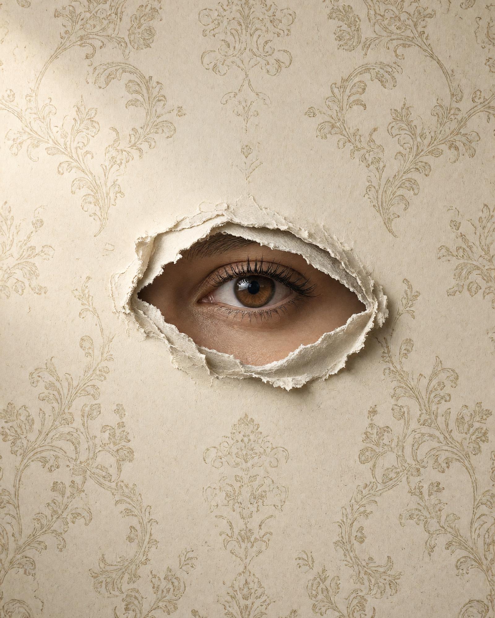 An eye looking out through a tear in ivory damask wallpaper